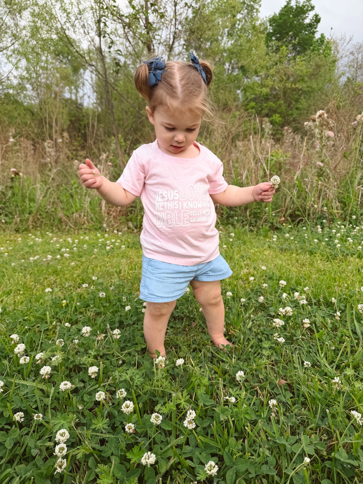 A little one exploring a clover field in a Jesus Loves Me tee